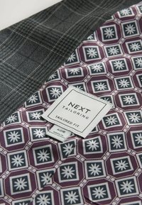 Tissu à carreaux gris avec une doublure géométrique violette, arborant une étiquette "NEXT Tailoring" et une étiquette de taille "40R". Texture lisse.
