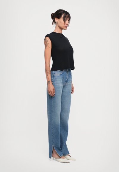 Black sleeveless top paired with high-waisted light blue wide-leg jeans featuring side slits. Model wears off-white heeled mules.