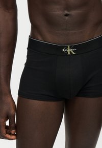 Άνδρας που φοράει μαύρα μποξεράκια Calvin Klein δείχνοντας τη μέση και το πάνω μέρος του μηρού σε απλό φόντο.