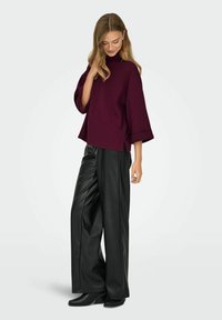Maglione oversize bordeaux con collo alto, maniche ampie e fessure laterali, abbinato a pantaloni a gamba larga in finta pelle nera.