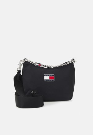 Zwarte Tommy Jeans schoudertas met zilveren kettingdetail, verstelbare stoffen riem en logopatch aan de voorkant tegen een effen achtergrond.