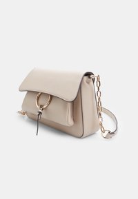 Borsa a tracolla in pelle beige con pattina pieghevole, hardware rotondo dorato e tracolla a catena. Presenta cuciture a contrasto e una tasca frontale.