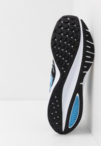 Scarpe da ginnastica in bianco e nero con un logo blu, inclinate su uno sfondo bianco, che mettono in mostra la suola testurizzata e il design dell'ammortizzazione.