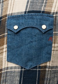 Poche boutonnée en denim bleu sur fond de tissu à carreaux marron et beige avec un petit détail brodé rouge.
