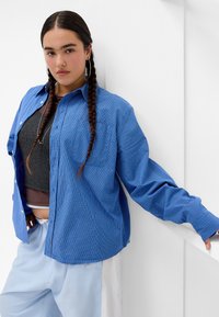 Blue checkered shirt with button closure, long sleeves, and a relaxed fit, layered over a gray cropped top. Light blue pants visible.