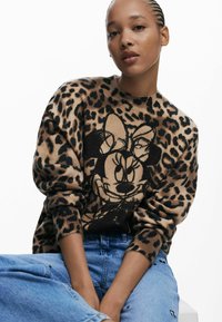 Pull en imprimé léopard beige avec des accents noirs, présentant un design de personnage de dessin animé. Texture douce et coupe décontractée avec des poignets côtelés.