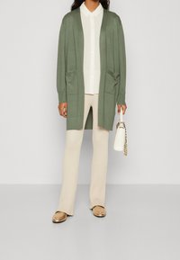 Kvinna som bär en lång olivgrön cardigan över en vit skjorta och krämfärgade ribbade byxor, håller en vit handväska med kedjereim.