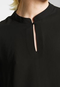 Gros plan sur le col d'une chemise noire avec un petit bouton rond et une ouverture verticale à l'avant, portée par une personne aux cheveux blonds.