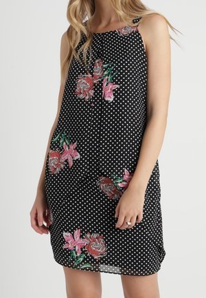 Kobieta w czarnej sukience bez rękawów w białe kropki i różowe kwiatowe wzory, stojąca na tle jednolitego jasnego tła.