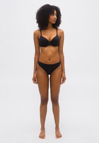 Γυναίκα με κατσαρά μαλλιά, όρθια και ξυπόλυτη, φοράει μαύρο σουτιέν και μαύρο εσώρουχο μπικίνι, κοιτάζοντας ελαφρώς προς τα δεξιά της.