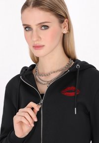 Sudadera con cremallera negra hecha de una tela suave, con un diseño de labios rojos en el pecho, cadenas plateadas en capas visibles, y un detalle de cremallera en la parte delantera.