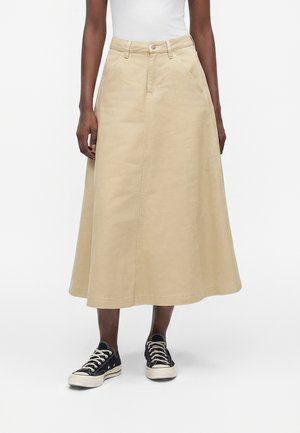 Γυναίκα φοράει μπεζ midi φούστα σε σχήμα Α με πλαϊνές τσέπες και μαύρα χαμηλά αθλητικά παπούτσια, στέκεται μπροστά σε μια απλή φόντο.