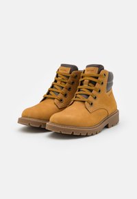 Botas de ante marrón hasta el tobillo con punta redonda, cordones amarillos y un collar acolchado. La suela de goma texturizada presenta profundas ranuras para un mejor agarre.