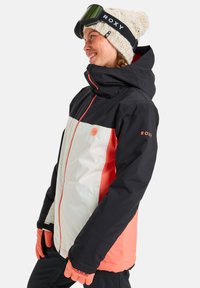 Giacca da sci bianca e nera con accenti corallo. Caratterizzata da un colletto alto, chiusura con zip e logo Roxy sulla manica. Tessuto strutturato.