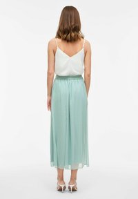 Světle zelená plisovaná maxi sukně s elastickým pasem, párovaná s bílým topem na tenkých ramínkách. Měkká, lehce splývavá látka s elegantním designem.