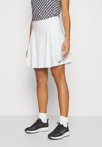 Bílá plisovaná sukně s širokým pasem, na kterém je malé černé logo Nike. Nošena s černými teniskami a bílými kotníkovými ponožkami.