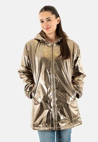 Jongeman met lang bruin haar, gekleed in een glanzende metalen gouden hoodie en blauwe jeans, met handen in de zakken, licht glimlachend.