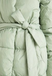 Manteau matelassé vert clair avec une taille ceinturée, une texture douce et un motif matelassé, agrémenté d'un nœud à l'avant.