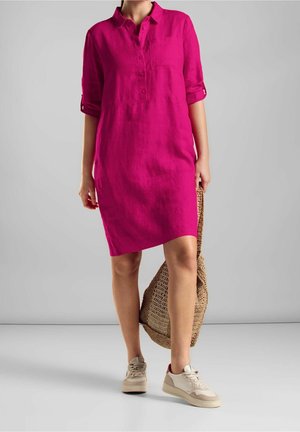 Osoba ubrana w jasnoróżową, sięgającą kolan sukienkę zapinaną na guziki z podwiniętymi rękawami, beżowe trampki i trzymająca plecioną torbę typu tote.