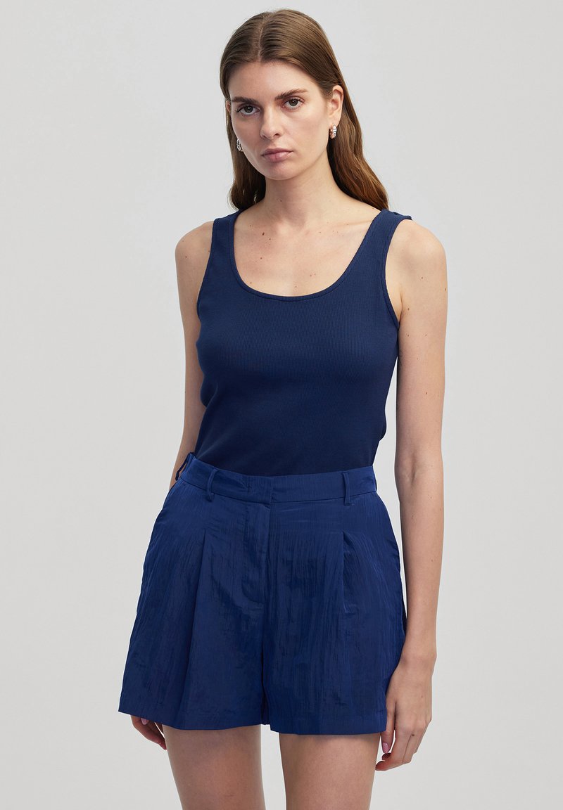 Marineblauwe geribde tanktop gecombineerd met geplisseerde marineblauwe shorts van een lichte, gestructureerde stof. Kenmerken zijn een ronde halslijn en een op maat gemaakte tailleband.
