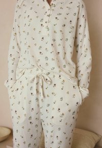Loungewear-Set in Creme mit floralem Muster aus strukturiertem Stoff. Das Oberteil hat einen geknöpften Ausschnitt, und die Hose verfügt über einen verstellbaren Bindegürtel an der Taille.