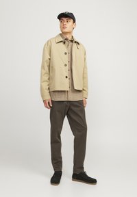 Beige cropped jacket with buttons, plaid shirt underneath, olive green pants, and black shoes, set against a neutral background.