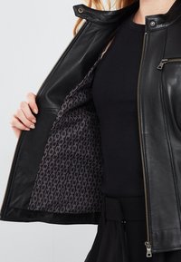 Black leather jacket with a zip closure, featuring a chain pattern lining. The exterior is smooth, with a snap collar and fitted design.