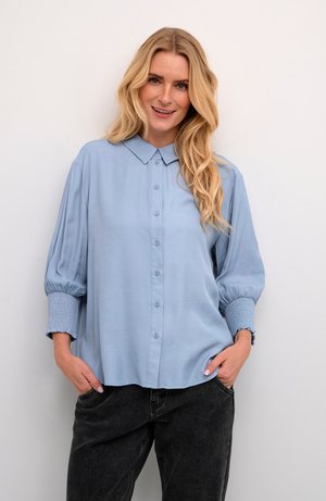 Lichtblauwe blouse met een knoopsluiting aan de voorkant, een kraag en opgezette mouwen met geraffineerde manchetten. Draagt met donkere, losvallende broeken.