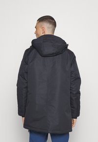 Svart, huva parkas med en slät, strukturerad tyg. Har en rak klippning och minimalistisk design, som slutar strax ovanför knäna.
