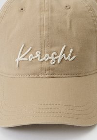 Beige színű pamut sapka ívelt karimával és fehér hímzett "Koroshi" logóval. Texturált anyag, látható varrási részletekkel.