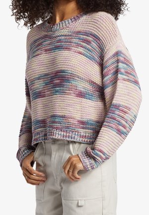 Femme portant un pull en tricot multicolore aux tons rose, bleu et violet, et un pantalon beige clair taille haute avec des poches.
