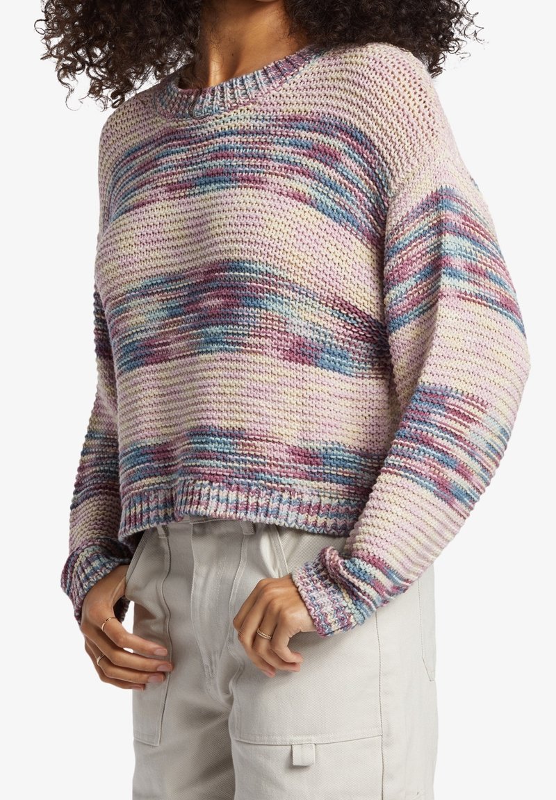 Femme portant un pull en tricot multicolore aux tons rose, bleu et violet, et un pantalon beige clair taille haute avec des poches.