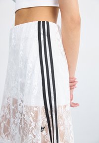 White lace skirt featuring black three-stripe accents, sheer texture, and an embroidered logo. Designed with a flowy, layered look.