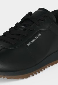 Baskets en cuir noir avec lacets noirs, semelle marron rainurée et marquage "MICHAEL KORS" sur le côté.