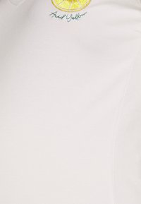 Camisa polo branca de algodão com um gráfico bordado de limão amarelo e texto cursivo verde na área do peito esquerdo.