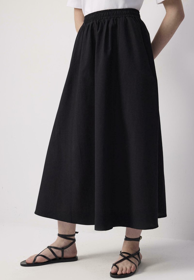 Persoană purtând o fustă neagră, evazată, de lungime până la gleznă, cu talie elastică și sandale negre plate cu barete, pe un fundal simplu.
