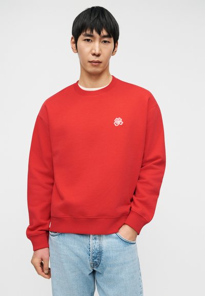 Άνδρας στέκεται με το ένα χέρι στην τσέπη, φοράει κόκκινο φούτερ με μικρό λευκό έμβλημα και ανοιχτόχρωμο τζιν μπροστά σε απλό φόντο.