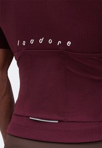 Κοντινή λήψη της πίσω τσέπης από βυσσινί ποδηλατική φανέλα με υφή, με το λογότυπο "Isadore" σε λευκά γράμματα πάνω από την τσέπη.