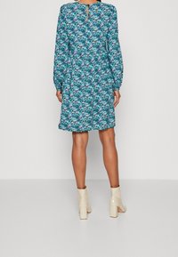 Blommig klänning med ett blått och grönt mönster, långärmad och med ett nyckelhål i ryggen, samt matchade beige klackankelstövlar.