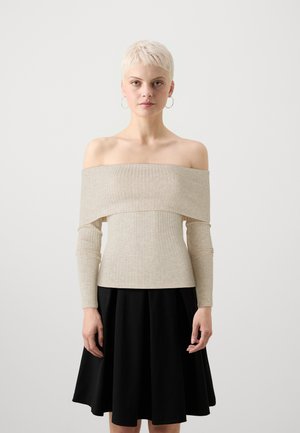 Top beige a spalla scoperta con texture a coste e maniche lunghe, abbinato a una gonna nera a campana. Lo sfondo è semplice e chiaro.