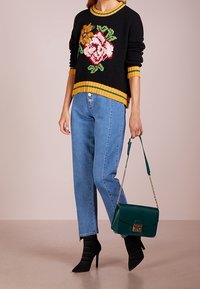 Černý pletený svetr s květinovým vzorem, žlutými a zelenými ribbovanými akcenty; v kombinaci se světle modrými džínami rovného střihu a zelenou kabelkou.