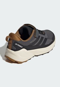 Chaussure de sport grise avec une tige en mesh noire, des accents marron, une semelle en caoutchouc texturée et une sangle réglable. Présente la marque TERREX.