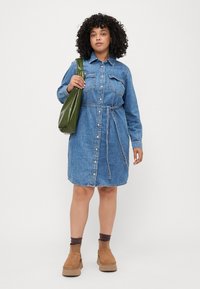 Denim skjortekjole med lange ermer, knappelukking, sidelommer og belte i stoff. Bar over en grønn veske og brune ankelstøvler.