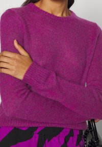 Fuchsia stickad tröja med långa ärmar, rund halsringning, räfflade muddar och en mjuk, texturerad yta. Stylar med en mönstrad kjol.