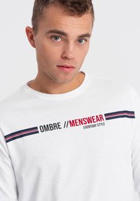 Weißes Langarmhemd aus Baumwolle mit schwarzen, roten und marineblauen Streifen, versehen mit dem Schriftzug "OMBRE // MENSWEAR" in fetter Schrift über der Brust.