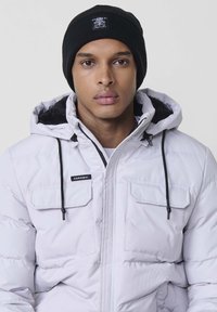 Giacca imbottita bianca con cappuccio e cordini neri, dotata di due tasche frontali e una toppa con logo. Berretto a maglia nero sulla testa del modello.