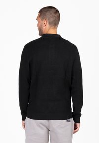 Schwarzer Strickpullover mit einer strukturierten Oberfläche. Mit Rundhalsausschnitt und langen Ärmeln. Der Rücken zeigt ein kleines Markenetikett am Saum.
