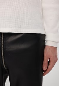 Hvid ribbet langærmet top med manchetter, parret med slanke sorte læderbukser, der har en synlig sølvlynlås detalje.