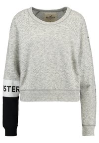 Sudadera gris de corte cropped con un acabado texturizado, que presenta una manga negra y una banda blanca con el logo "HOLLISTER". Puños y cuello acanalados.