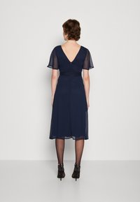 Marinblå klänning med korta, genomskinliga puffärmar, V-format rygg och en figursydd midja. Kjolen är knälång med en lätt utsvängd design.